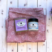 Load image into Gallery viewer, Two products from Heritage Homestead on a pink heart-patterned towel with a wooden background and lavender flowers.