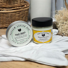 Load image into Gallery viewer, Two containers of body care products on a wooden surface with a white cloth and wicker basket in the background.