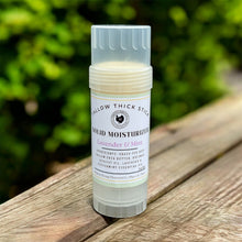 Load image into Gallery viewer, A single solid moisturizer stick in a clear cylindrical container with a white label reading 'TALLOW THICK STICK' and 'SOLID MOISTURIZER Lavender & Mint,' standing on a wooden surface with blurred green foliage in the background.