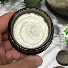 Load image into Gallery viewer, A hand holding an open round amber glass jar filled with a thick white cream or balm, photographed close up against a light surface with soft shadows and small green leaves and stones in the background.