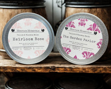 Load image into Gallery viewer, Two tins of Heritage Homestead candles labeled 'Heirloom Rose' and 'The Garden Parlor' on a wooden surface.
