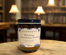 Load image into Gallery viewer, dickens Christmas night tallow and bees wax candle on a wooden surface with a blurred library background with old books on shelves and lights. 
