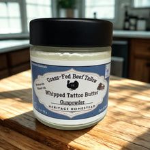 Load image into Gallery viewer, Heritage homestead Whipped tallow tattoo butter in a 3oz glass jar with black lid on a natural wooden counter with a blurred kitchen background. 
