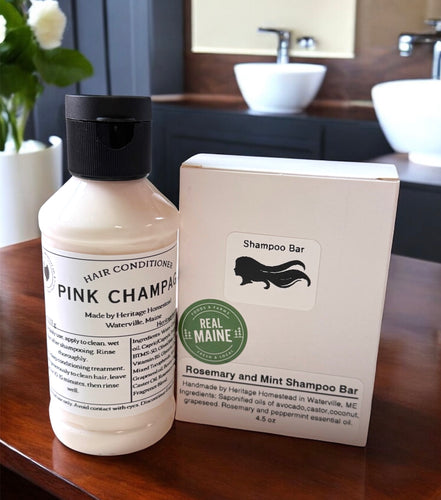 A natural hair care duo displayed on a polished wooden countertop in a modern bathroom setting. On the left is a bottle of Pink Champagne Hair Conditioner made with nourishing beef collagen, housed in a creamy pink formula with a black flip-top cap. On the right is a Rosemary and Mint Shampoo Bar in minimalist white packaging, featuring a silhouette of flowing hair and a green 