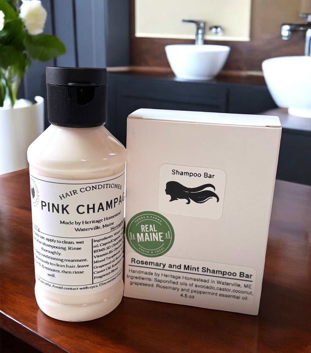 A natural hair care duo displayed on a polished wooden countertop in a modern bathroom setting. On the left is a bottle of Pink Champagne Hair Conditioner made with nourishing beef collagen, housed in a creamy pink formula with a black flip-top cap. On the right is a Rosemary and Mint Shampoo Bar in minimalist white packaging, featuring a silhouette of flowing hair and a green 