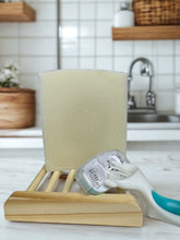 Load image into Gallery viewer, Unscented handcrafted tallow shave bar displayed on a wooden soap dish beside a multi-blade razor, with a bright farmhouse-style bathroom blurred in the background.
