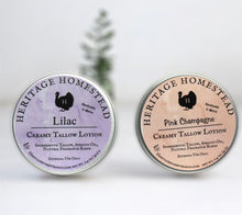 Load image into Gallery viewer, Two round tins of creamy tallow lotion from Heritage Homestead are displayed on a white surface with a soft-focus evergreen tree in the background. The left tin is labeled &quot;Lilac&quot; with a purple watercolor background, and the right tin is labeled &quot;Pink Champagne&quot; with a blush-pink background. Both tins feature the Heritage Homestead logo with a silhouette of a bird and include ingredients such as tallow, apricot oil, and natural fragrance blend.
