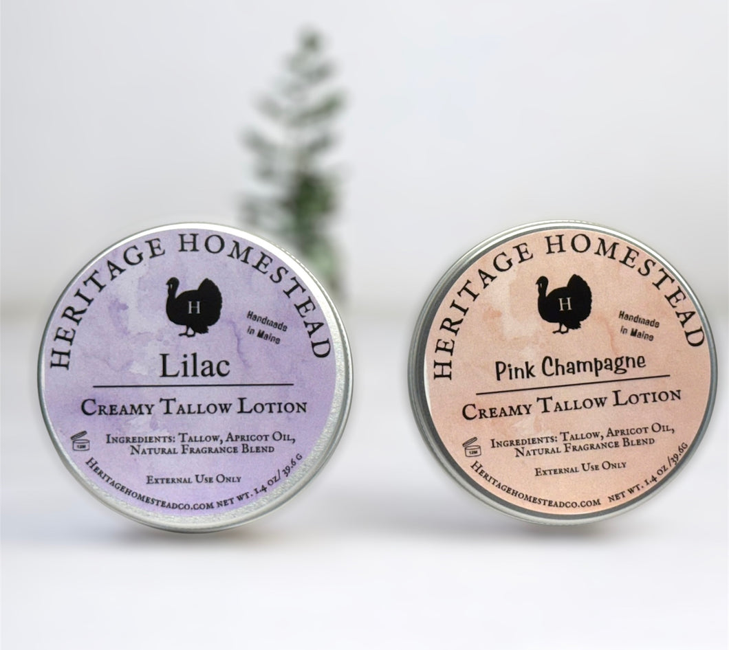 Two round tins of creamy tallow lotion from Heritage Homestead are displayed on a white surface with a soft-focus evergreen tree in the background. The left tin is labeled 