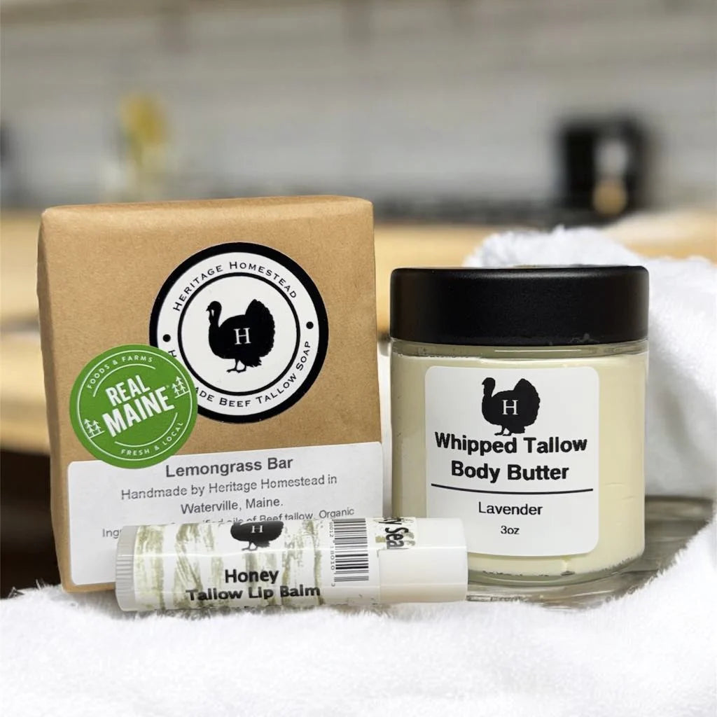 Natural tallow skincare gift set including a Lemongrass Bar soap, Whipped Tallow Body Butter in lavender scent, and a Honey Tallow Lip Balm. All products are handcrafted by Heritage Homestead in Waterville, Maine, and displayed on a soft white towel with a kitchen background.
